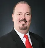 A smiling man with short hair and a goatee wearing a black suit, white shirt, and red tie. He is posed against a plain dark background.