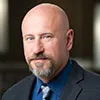 A headshot of a bald man with a trimmed beard, dressed in a dark suit, blue shirt, and patterned tie. He has a neutral expression and is photographed against a softly blurred indoor background.