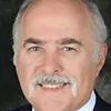 A headshot of a man with a distinctive mustache and bald head. He is wearing a suit jacket and tie, smiling slightly, and is set against a dark professional background.