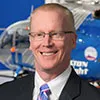 A professional headshot of a man with short, light red hair and glasses. He is wearing a dark suit, a light blue shirt, and a striped tie.