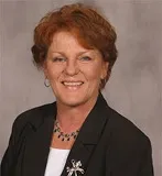 A smiling woman with short red hair wearing a black blazer over a white top, accessorized with a silver brooch and necklace. She is posed against a plain gray background.