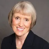 A professional headshot of a woman with short, light blonde hair styled neatly in a classic bob. She is smiling warmly and wearing a dark blazer with a neutral background, projecting a polished and approachable demeanor.