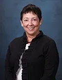 A portrait of a woman with short, dark hair and light skin. She is smiling and wearing a black jacket with a ruffled collar over a white top. The background is a textured blue, giving the image a formal and polished appearance.