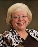 A professional headshot of a smiling woman with short, light blonde hair. She is wearing glasses, a gold chain necklace, and a patterned brown and white top over a dark blouse.