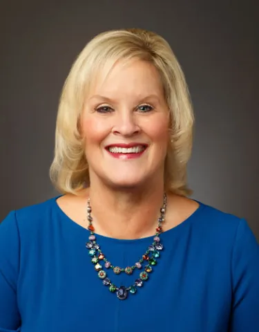 A professional headshot of a woman with shoulder-length blonde hair, wearing a vibrant blue blouse and a multi-colored statement necklace.