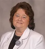 A woman with shoulder-length dark brown hair is wearing a white blazer with black piping and a dark blouse underneath. She is smiling softly, and a large decorative brooch is pinned near her collar.