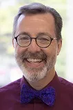 A smiling man with short gray hair and a beard, wearing round glasses and a burgundy shirt with a vibrant blue bow tie. The background appears softly blurred, suggesting an indoor setting with natural light.