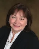 A smiling woman with shoulder-length dark brown hair wearing a white blouse and a black blazer, accessorized with small hoop earrings. She is posed against a softly blurred dark background.