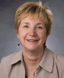 A smiling woman with short blonde hair wearing a beige blazer over a light top, accessorized with drop earrings and a silver necklace. She is posed against a softly blurred gray background.