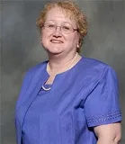 Portrait of a woman with short curly blonde hair, wearing glasses, a blue blouse, and a necklace, smiling softly.