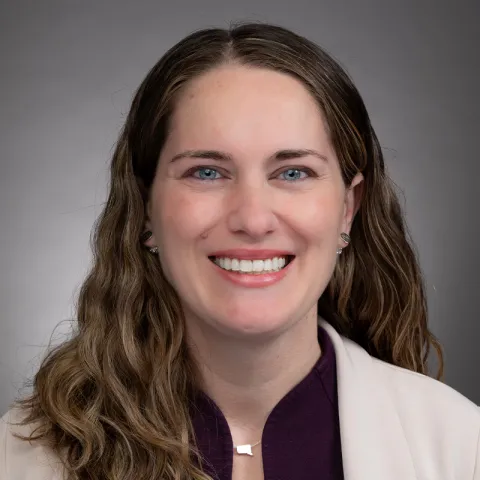 A portrait of a woman with wavy, medium-length light brown hair and light skin. She is smiling brightly and wearing a purple blouse with a light beige blazer. She has small silver earrings and a minimalist silver necklace. The background is a neutral gray gradient, giving the image a professional and polished appearance. Her expression is friendly and approachable.