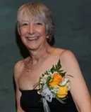 A smiling woman with short gray hair wearing a strapless black dress and a floral corsage featuring yellow and white flowers with greenery. She is posed against a dark green textured background.