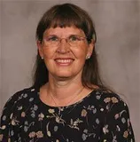 A professional portrait of a woman with long, straight brown hair and bangs. She is wearing glasses and a patterned blouse with floral designs. The background is a soft, light gray tone.