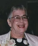 A smiling older woman with short gray hair wearing glasses. She is dressed in a white jacket over a polka-dot blouse, with a floral corsage pinned to her attire. The background is softly blurred.