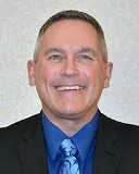 A smiling man with short gray hair wearing a black suit, blue shirt, and a blue patterned tie. He is posed against a light textured background.