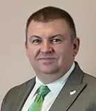 A professional headshot of a man wearing a gray suit, white shirt, and green tie, with a small lapel pin on his suit jacket. He has short brown hair and a neutral expression, set against a light beige background.