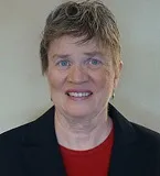 A woman with short gray hair wearing a black blazer over a red top. She has a neutral expression and is posed against a plain light-colored background.