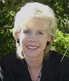 A smiling woman with short blonde hair wearing a black blazer and black earrings. She is posed outdoors with greenery in the background.