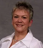 A smiling woman with short spiked gray hair wearing a white collared shirt, drop earrings, and a silver necklace. She is posed against a plain gray background.