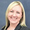 A portrait of a woman with straight, shoulder-length blonde hair and light skin. She is smiling warmly and wearing a dark blazer over a light top. The background is a solid muted blue, creating a professional and polished appearance. Her expression is friendly and approachable.