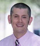 A smiling man wearing a light pink collared shirt and a striped tie in shades of black, white, and pink. The background is softly blurred with outdoor lighting.