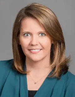 Headshot of a woman with shoulder-length blonde hair, wearing a teal blazer and a necklace, smiling warmly.