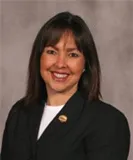 A portrait of a woman with shoulder-length dark brown hair and light skin. She is smiling warmly and wearing a black blazer over a white top, with a small gold pin visible on her blazer. 