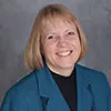 A professional portrait of a woman with short blonde hair, smiling warmly. She is wearing a teal blazer over a black top. The background is a neutral gray, complementing her professional attire.