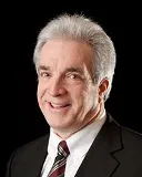 A smiling man with short gray hair wearing a black suit, white shirt, and striped tie. He is posed against a plain black background.