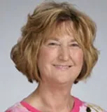 A portrait of a woman with shoulder-length, wavy blonde hair and light skin. She is smiling gently and wearing a pink top with a patterned design.