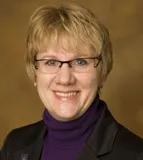 A professional portrait of a woman with short, blonde hair wearing glasses. She is dressed in a black blazer over a purple turtleneck sweater. The background is a warm, gold-toned gradient.