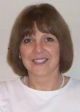 A smiling woman with shoulder-length brown hair and bangs wearing a white top and a pearl necklace. She is posed against a plain light background.