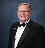 A professionally captured photo of a man in formal attire, wearing a black tuxedo and a bow tie. The subject is smiling confidently against a blue studio background.