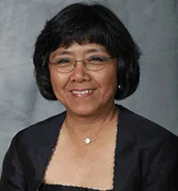 A smiling woman with short black hair and glasses wearing a black formal outfit with a necklace featuring a small circular pendant. She is posed against a plain gray background.