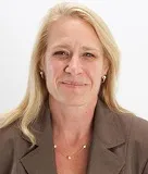 A woman with long blonde hair wearing a brown blazer and a delicate necklace. She has a neutral expression and is posed against a plain white background.