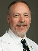 A smiling man with short gray hair and a trimmed beard wearing a white lab coat, a striped shirt, and a striped tie. He is posed against a plain dark background.