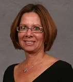 A smiling woman with shoulder-length brown hair and glasses wearing a black top, accessorized with a silver necklace. She is posed against a plain gray background.