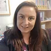 A professional portrait of a woman with long brown hair, wearing a black blazer and a patterned top. She is smiling warmly in an office setting, with shelves and framed pictures visible in the softly blurred background, creating a friendly and approachable impression.