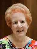 A smiling woman with short, styled reddish hair wearing a colorful floral-patterned top. She is posed against a softly blurred wooden background.