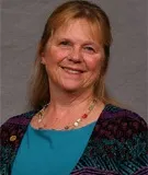 A smiling woman with long blonde hair tied back wearing a colorful patterned cardigan over a teal top, accessorized with a multicolored necklace and a small lapel pin. She is posed against a plain gray background.