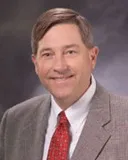 A portrait of a man with short, neatly combed brown hair and light skin. He is smiling warmly and wearing a gray suit, a white dress shirt, and a red tie.