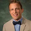 A professional portrait of a man with short, light brown hair styled neatly. He is dressed in a light beige suit jacket, a blue shirt with subtle patterns, and a dark blue bow tie with white accents.