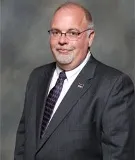 A professional portrait of a man with short, light-colored hair and glasses, dressed in a dark suit, white shirt, and a patterned tie. The background is neutral and blurred, adding a formal and polished look to the image.