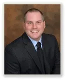 A smiling man with short light brown hair wearing a dark gray suit, light blue shirt, and black tie. He is posed against a softly blurred brown background.