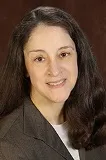 A portrait of a woman with long, dark brown hair and fair skin. She is wearing a gray blazer over a white top and has a gentle smile. The background is a warm brown tone, giving the image a professional and composed appearance. Her expression is approachable and kind.