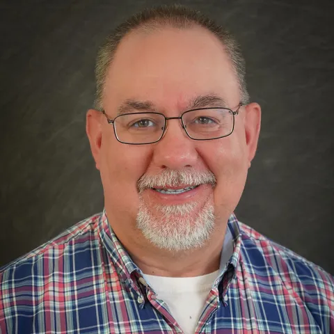 Professional headshot of a smiling middle-aged man with glasses, a white beard, and a plaid shirt, set against a dark textured background.