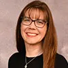 Headshot of a woman with long brown hair and bangs, wearing glasses, a black top, and a silver necklace, smiling warmly.