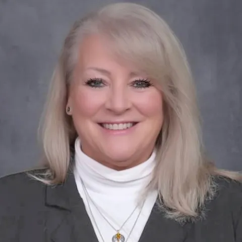 Professional headshot of a smiling woman with long blonde hair, wearing a dark blazer over a white turtleneck and layered necklaces, set against a gray backdrop.