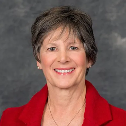 Professional headshot of a smiling woman with short dark hair, wearing a red blazer and a delicate necklace, set against a gray textured background.