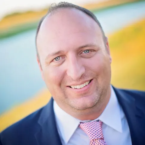 Professional headshot of a smiling man with short dark hair, wearing a navy blue suit, white shirt, and pink patterned tie, set outdoors with a blurred background of water and greenery.